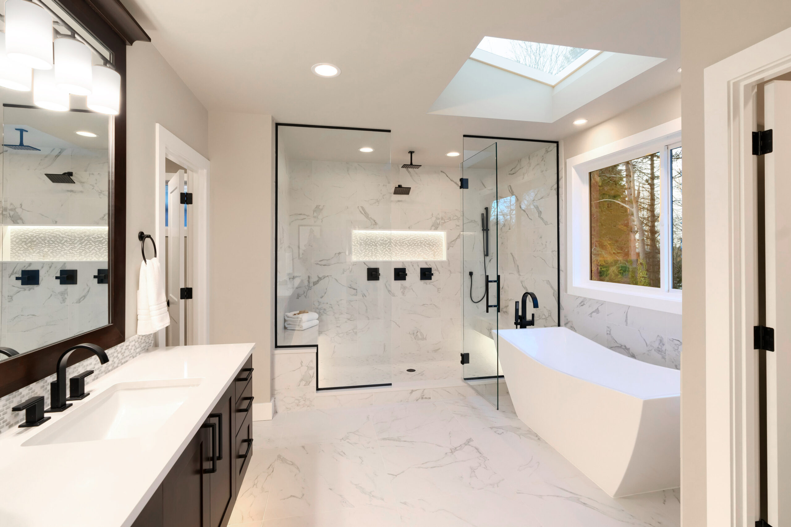 Luxury modern home bathroom interior with dark brown cabinets, white marble, walk in shower, free standing tub, two mirrors.