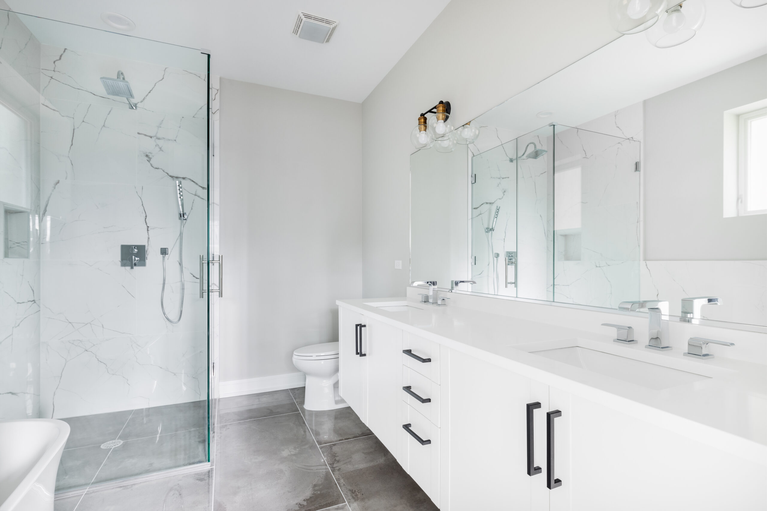 A modern, white luxury bathroom with black hardware and chrome faucets. A large glass walled shower sits in the corner with marble tiles from floor to ceiling.